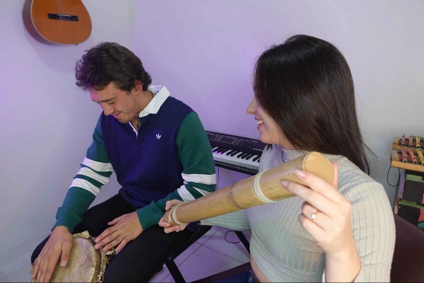 Two people sitting, one holding a drum, in a room with a keyboard and a guitar in the background. 5
