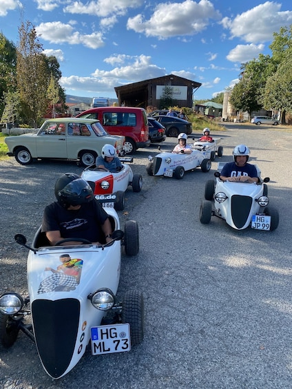 A group of people in go-karts on a gravel path with cars parked in the background. 4