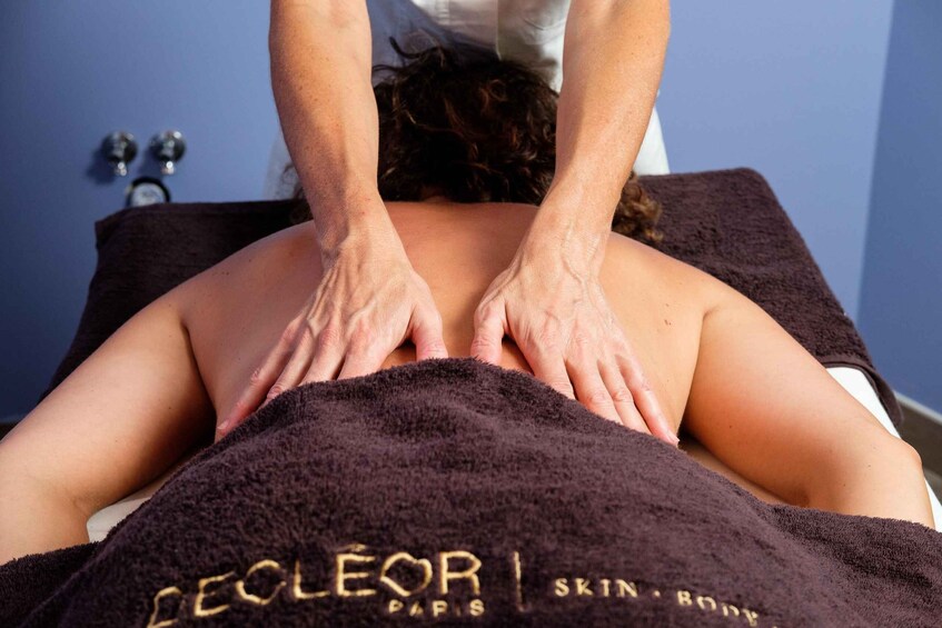 A person receiving a back massage with hands on their back, covered by a towel with 'DELEOR PARIS' text. 5