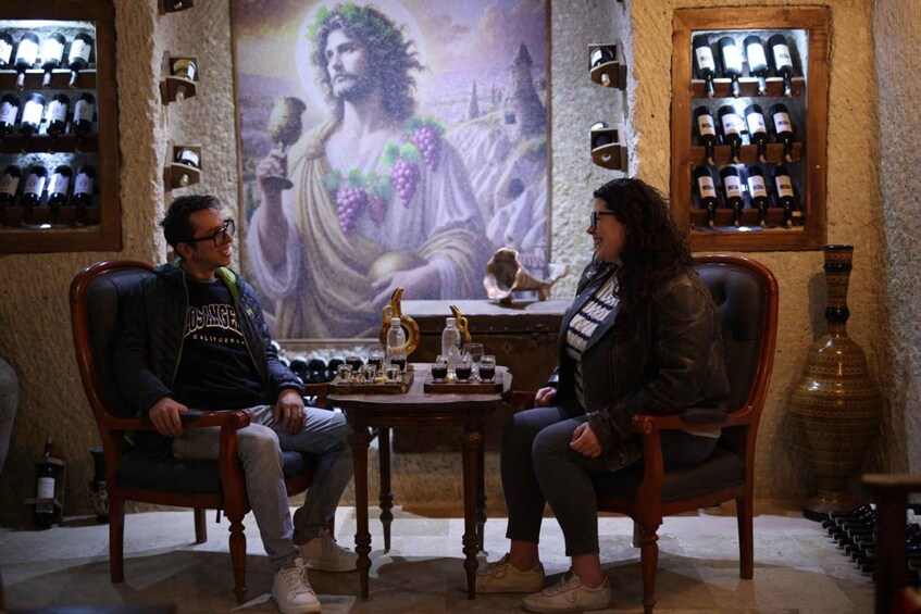 Two people seated at a table with wine bottles and glasses in a room with a large painting and wine racks. 3