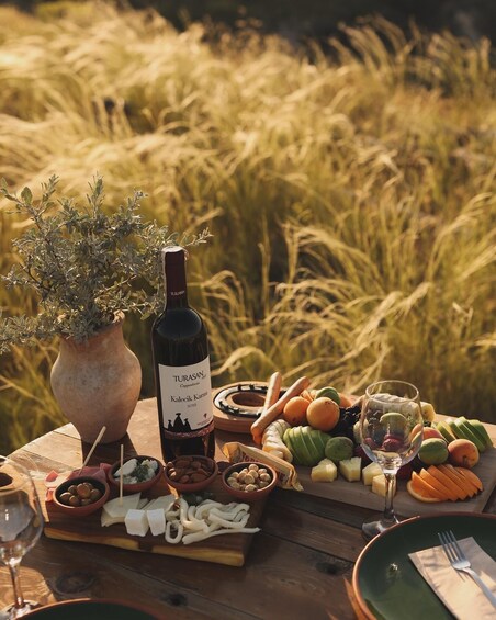 A picnic table with a bottle of wine, a vase with a plant, and a variety of fruits and cheeses. 2
