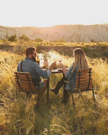 Two people sitting at a table outdoors with a bottle and glasses, surrounded by tall grass and a hilly landscape. 1