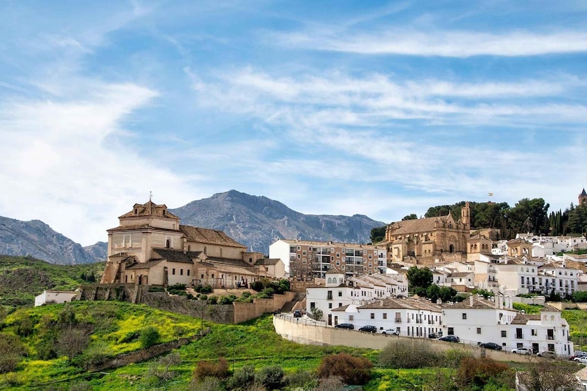 A historic town with white buildings, a prominent church, and mountains in the background. 1