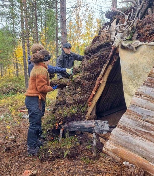 Three people engaged in clearing vegetation around a wooden structure in a forested area. 1