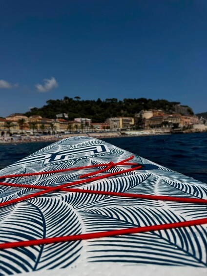 A boat's wake with a patterned sail and red ropes, heading towards a coastal town with buildings and trees. 4