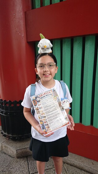 A child holding a booklet titled 'Asakusa Ninja Mission' with a plush toy on their head. 3