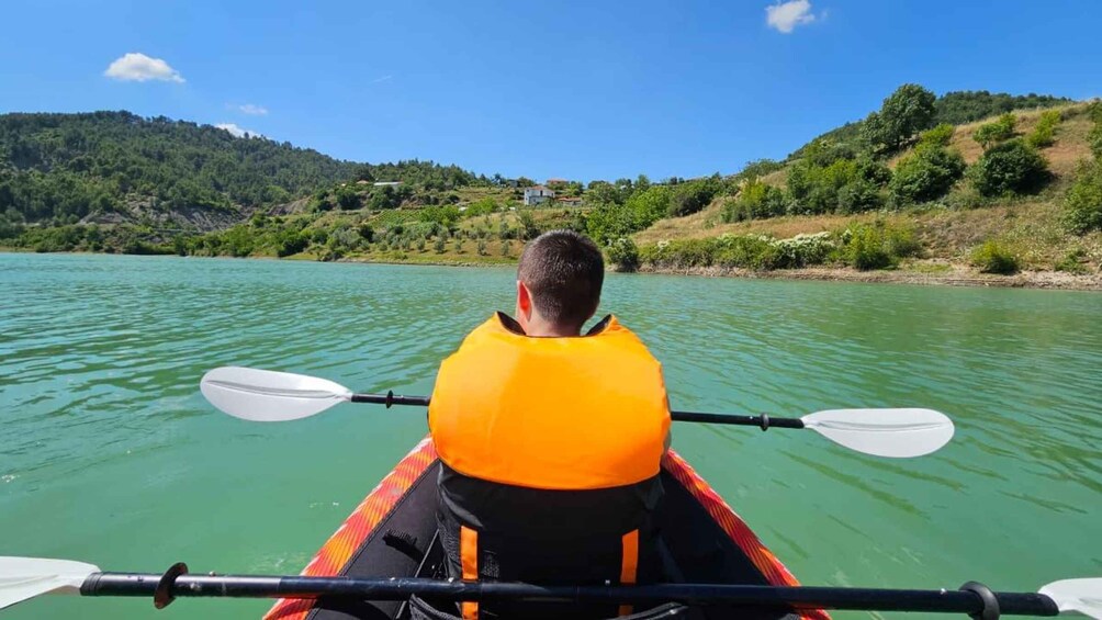 A person kayaking on a calm river with green water, surrounded by hills and sparse vegetation. 1