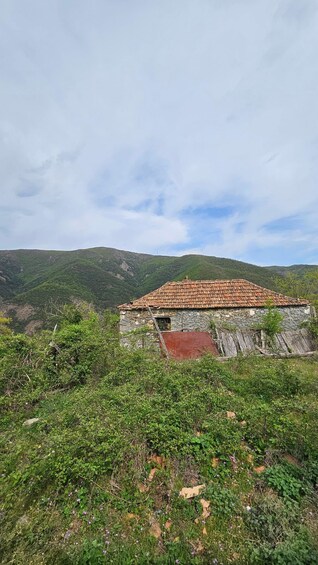 A rustic stone building with a tiled roof, surrounded by lush greenery and hills in the background. 3