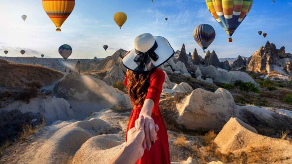 A person in a red dress holding hands with another, standing on rocky terrain with hot air balloons in the background. 1