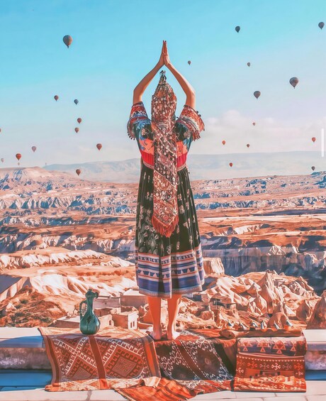 A person in traditional attire standing on a plateau with a mountainous landscape and hot air balloons in the sky. 3