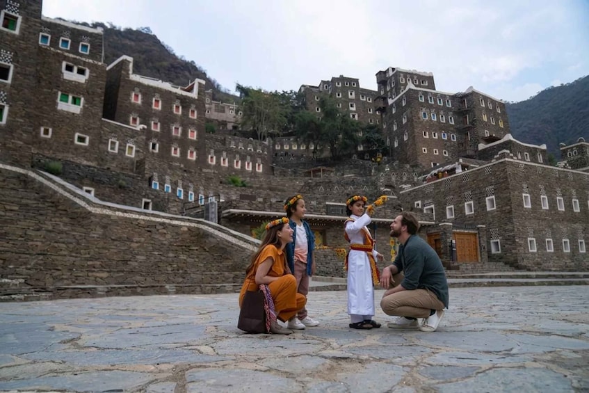 A group of people in traditional attire in front of a historic stone fortress with multiple windows and balconies. 2