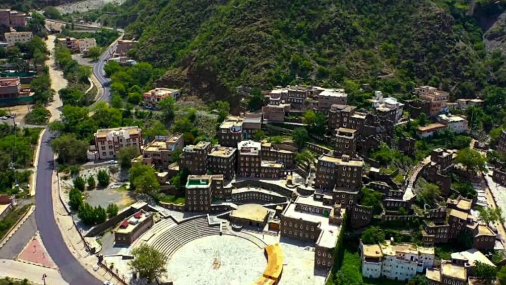 A densely built urban area with a central open space surrounded by buildings, a curved road to the left, and a mountainous backdrop. 3