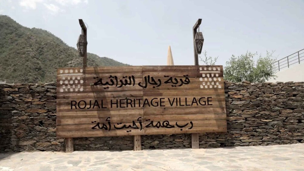 A wooden sign with 'ROJAL HERITAGE VILLAGE' written in English and another script, set against a stone wall and a mountainous backdrop. 5