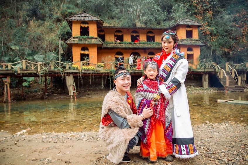 A family in traditional attire standing in front of a rustic building with a river in the background. 1