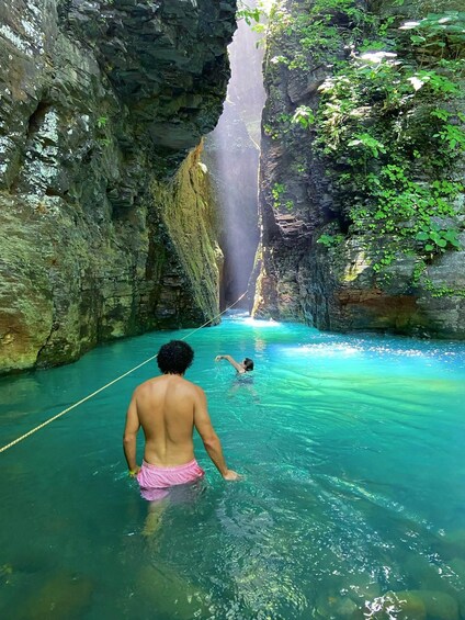 A man in a cave with a rope, another person swimming, and a waterfall in the background. 5