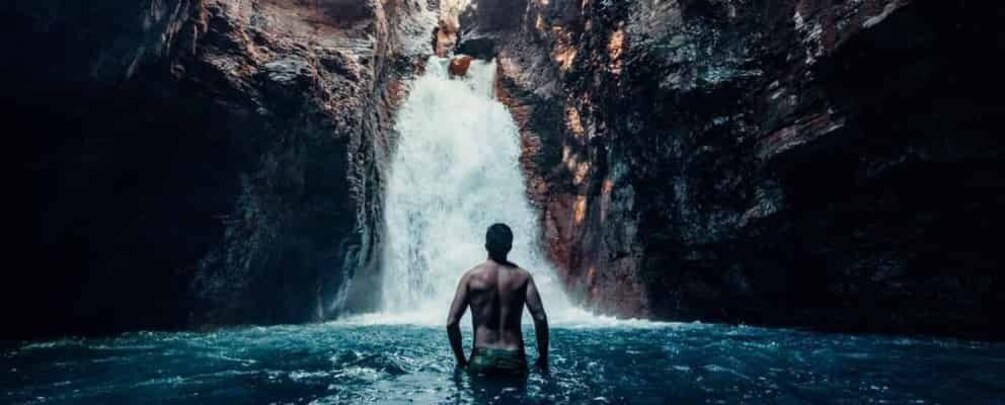 A person standing in water facing a waterfall in a rocky cave. 1