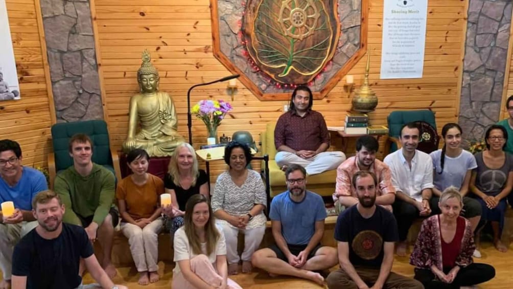 A group of people seated in a room with wooden walls, a large Buddha statue, and a decorative wall hanging. 2