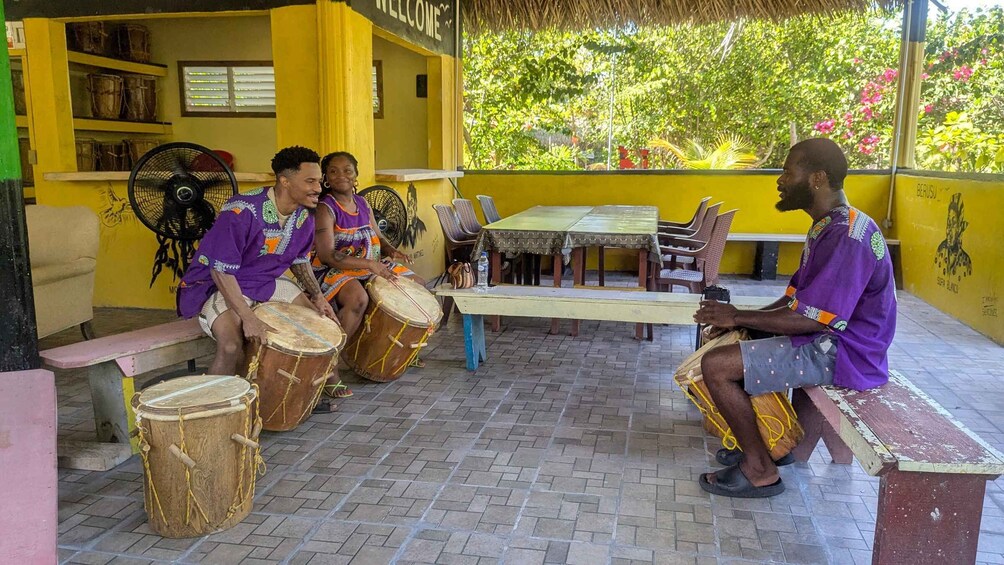 A group of people playing drums in an outdoor setting with a yellow building and a thatched roof in the background. 1