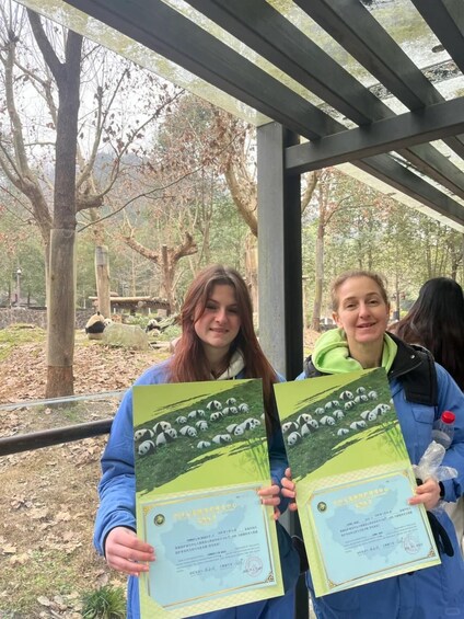 Two individuals holding certificates in front of a zoo enclosure with pandas. 1