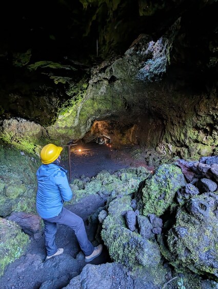 A person in a yellow hard hat and blue jacket exploring a cave with moss-covered rocks and a small pool of water. 3