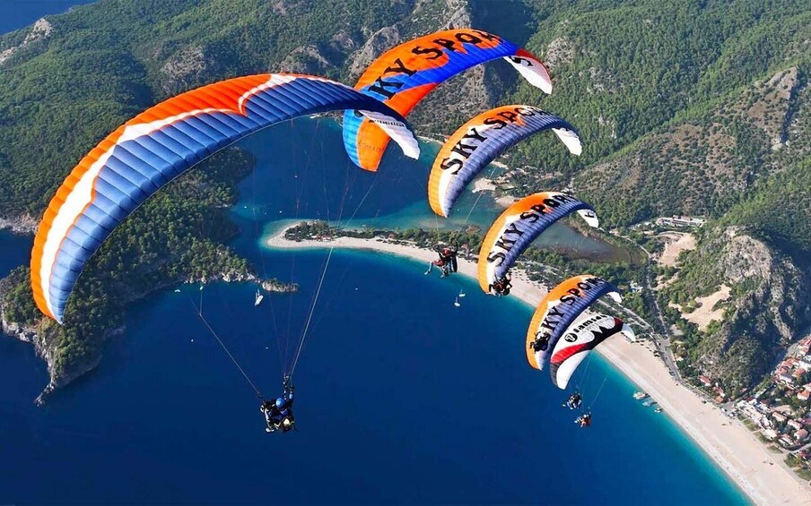 Paragliders with 'SKYSPORT' banners flying over a coastal area with a beach and forested hills. 4