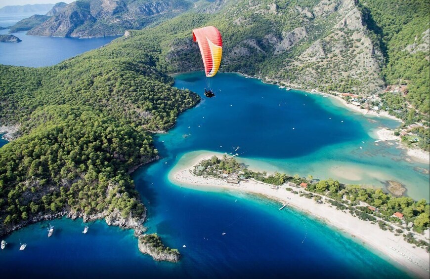 A paraglider over a coastal area with clear blue waters, green hills, and a small island with buildings. 2