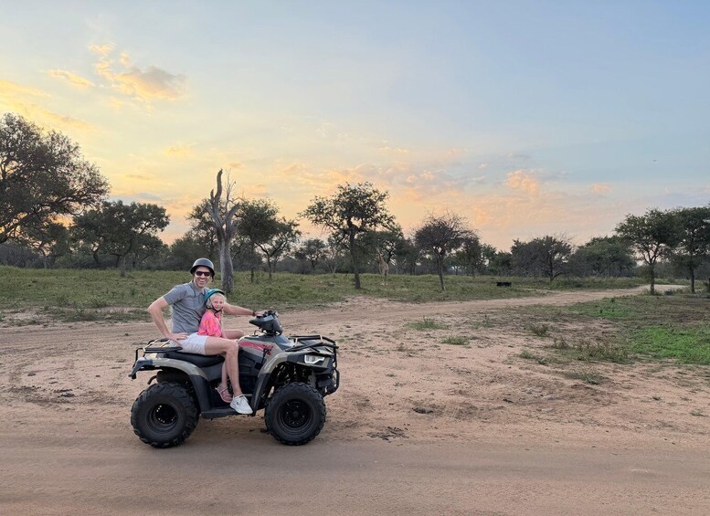 A man and a child on an ATV in a dry, open landscape with sparse trees and a clear sky. 2