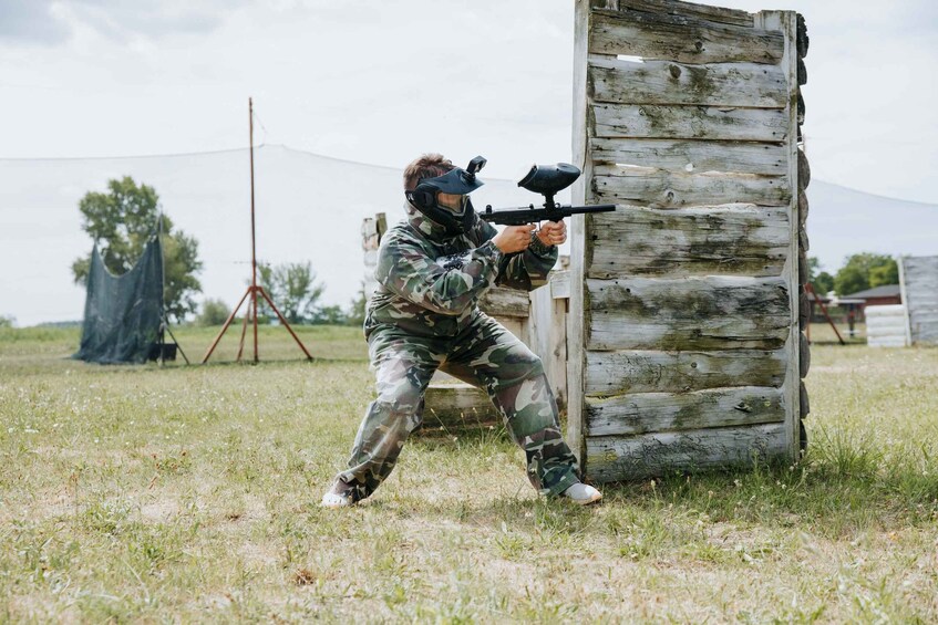 A person in camouflage paintball gear aiming a paintball gun at a wooden structure. 2