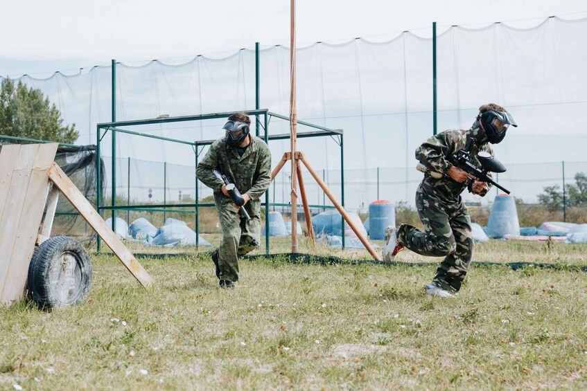 Two people in camouflage paintball gear, one aiming a paintball gun, and a paintball arena with a netted enclosure and wooden structures. 3