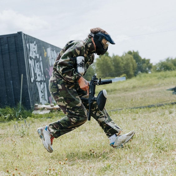 A person in camouflage gear running with a paintball gun, grass, and a building with graffiti in the background. 3