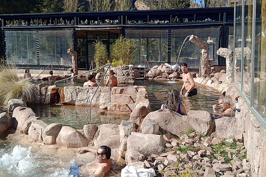 A natural hot spring area with people relaxing in the water, surrounded by rocks and a glass bridge overhead. 2