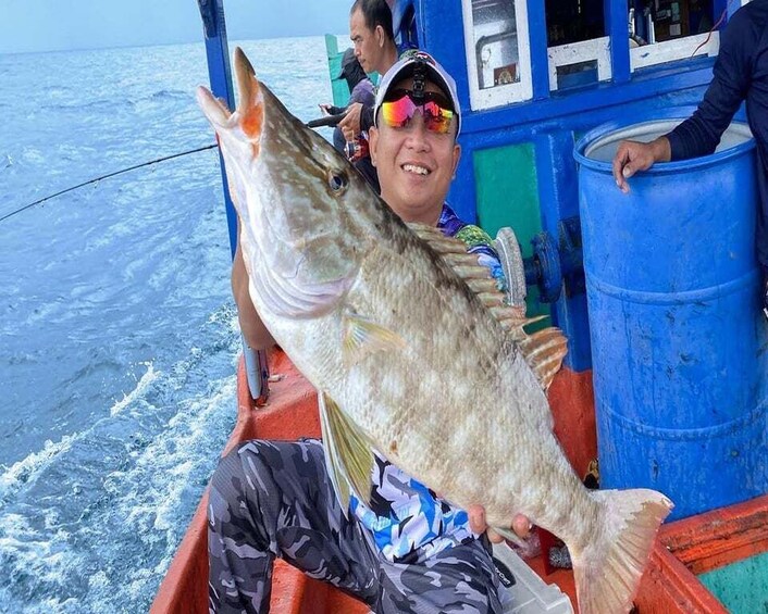 A person holding a large fish on a boat with a blue barrel and a person in the background. 2