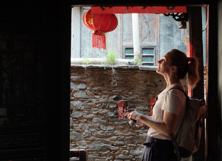 A person holding a camera, standing at a doorway with a stone wall and a red lantern above. 4