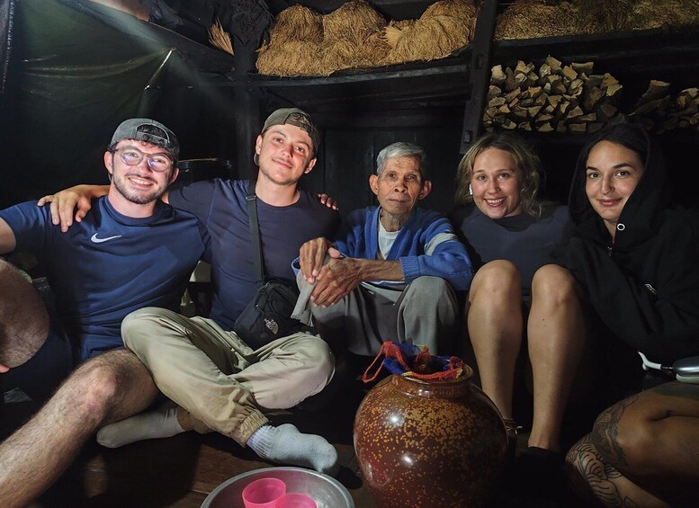 A group of people sitting indoors with a large pot and a stack of firewood in the background. 2