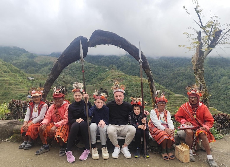 A group of people in traditional attire seated under a natural archway with a mountainous landscape in the background. 1