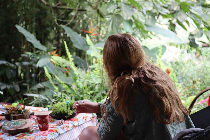 A person with long hair sitting at a table with a floral tablecloth, surrounded by greenery and plants. 3