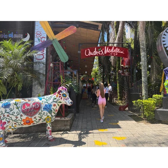 A colorful street scene with a large decorative cow statue, a sign for 'Audres Medellin,' and a clock tower in the background. 2