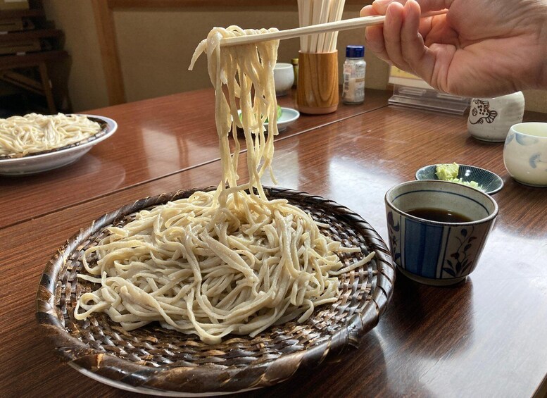 A bowl of noodles being lifted with chopsticks, a cup of tea, and a small dish of sauce on a wooden table. 4