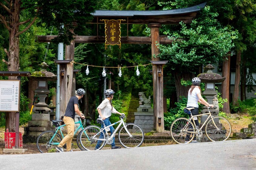 Three cyclists passing under a traditional Japanese torii gate surrounded by lush greenery. 1