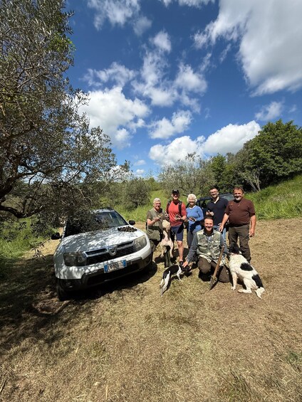 A group of people with a dog and a car in a grassy area with trees. 1