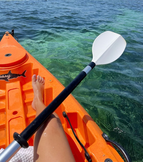 A person sitting on an orange kayak with a white paddle, feet dangling in the water. 3
