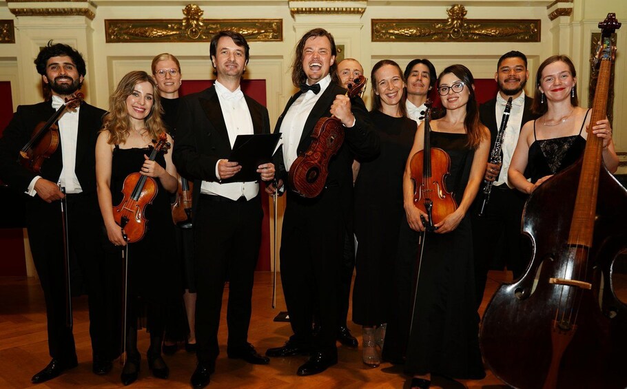 A group of musicians in formal attire holding string instruments in a room with ornate decorations. 2