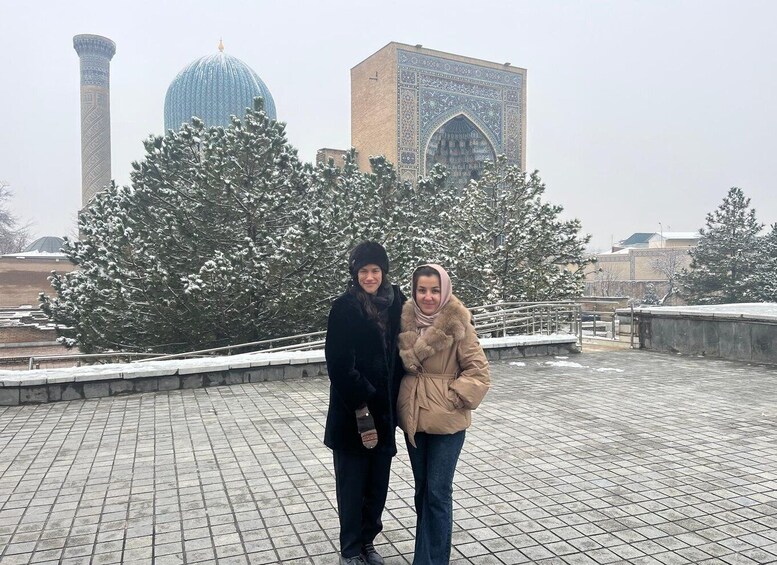 Two people standing on a tiled plaza with snow-covered trees and a large blue dome and minaret in the background. 4