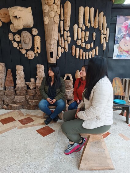 A group of people seated on stools in front of a wall displaying various wooden carvings. 5