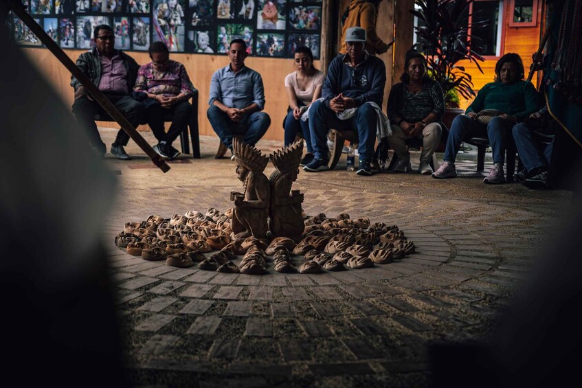 A group of people seated around a central sculpture on a cobblestone floor, with a wall of artwork in the background. 3