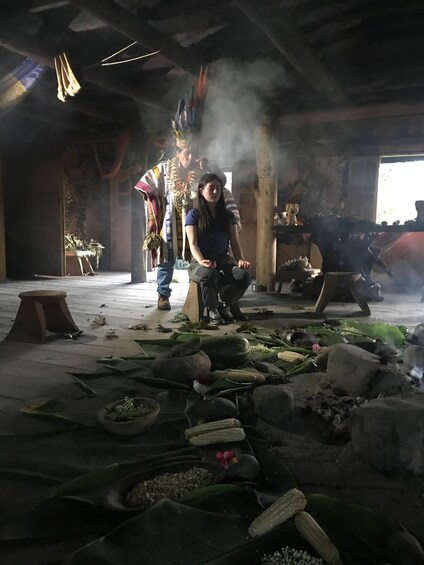 A person sitting on a stool in a dimly lit room with smoke, surrounded by scattered food items and a large plant. 1