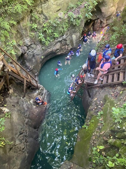 A group of people in water wings and helmets at a rocky water body with a wooden staircase. 1
