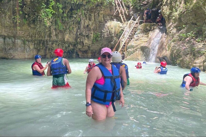 People in water with life vests and helmets, a waterfall, and rocky cliffs. 2