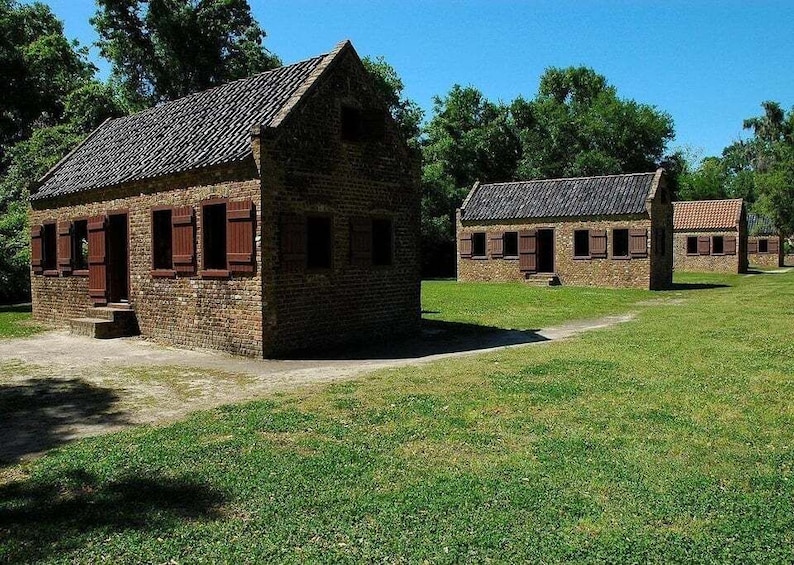 A row of stone buildings with wooden shutters, set in a grassy field with trees in the background. 4