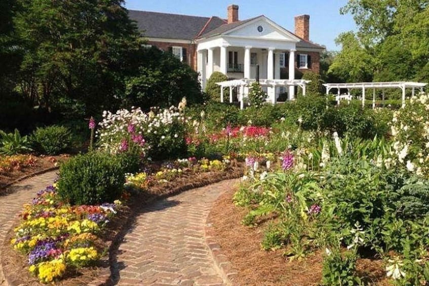 A garden with a brick pathway leading to a white house with columns and a porch. 5